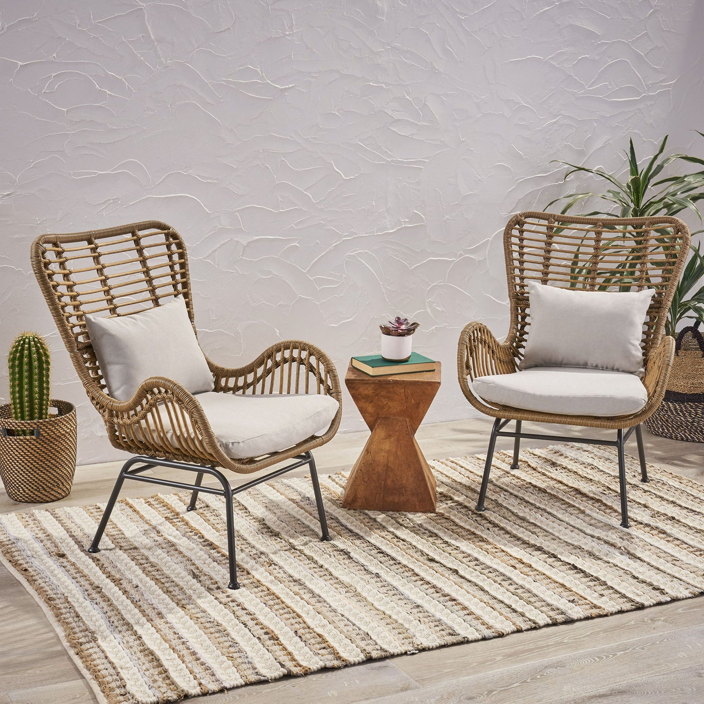 Two Modern Aesthetic Outdoor Rattan Accent Chairs with white cushions sit on a striped rug, facing a small wooden side table with a succulent. In the background, there’s a potted cactus and leafy plant against a textured white wall.