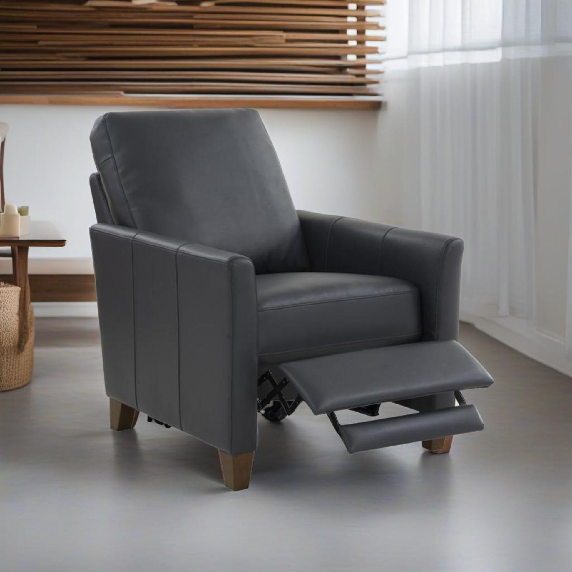 The Pablo - Modern Recliner With Manual Recline, featuring gray leather and wooden legs, sits in a bright room with sheer white curtains and wooden decor. Its footrest is extended for added comfort.
