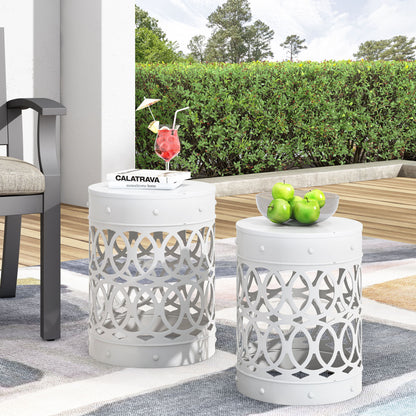 Two white Outdoor Metal S/2 End Tables with geometric cutouts sit on a patio—one holds a drink and book, the other a bowl of green apples. A chair, rug, and greenery are visible in the background.