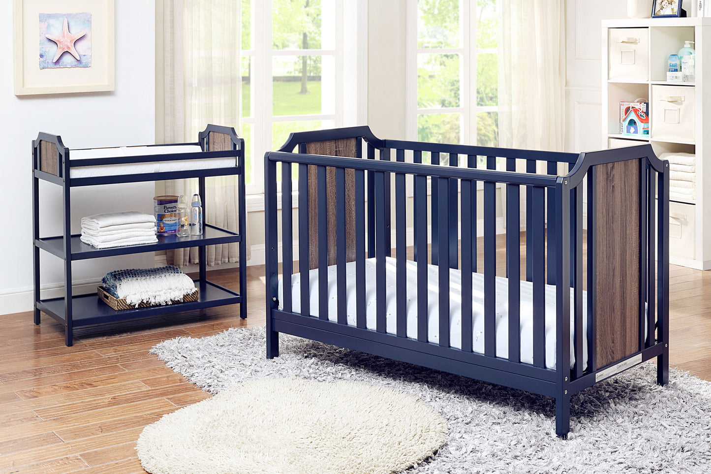 A bright nursery with large windows and wood floors features a navy blue crib and the Brees - Changing Table with shelves, a round rug, and baby supplies. Natural light fills the room.