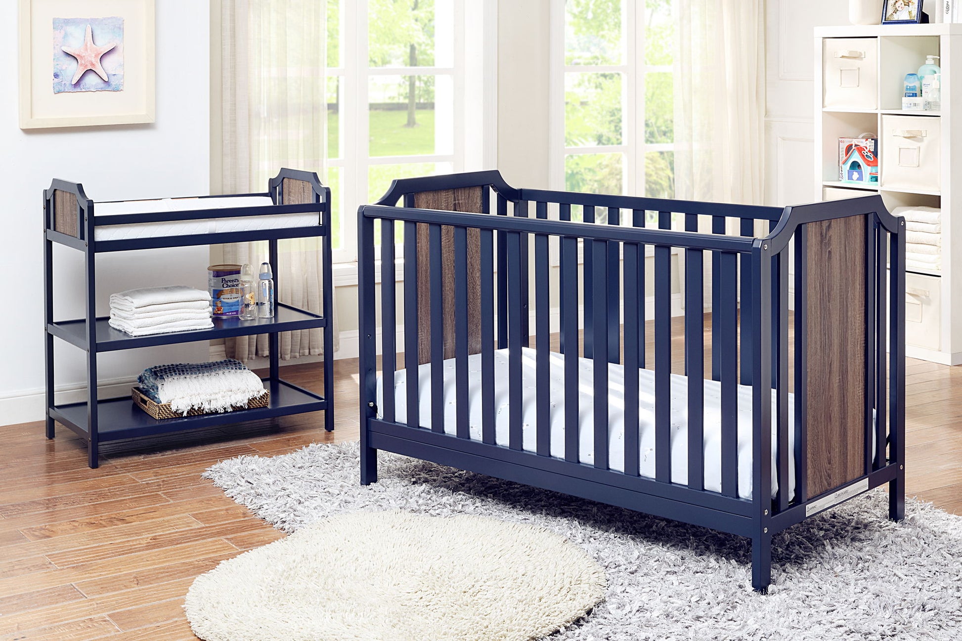 A bright nursery with large windows and wood floors features a navy blue crib and the Brees - Changing Table with shelves, a round rug, and baby supplies. Natural light fills the room.