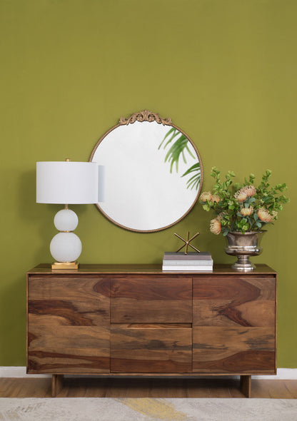 A wooden sideboard stands against an olive green wall, topped with a white lamp, books, a sculpture, silver vase with flowers, and the Classic Design Mirror With Baroque Inspired Frame leaning behind them.
