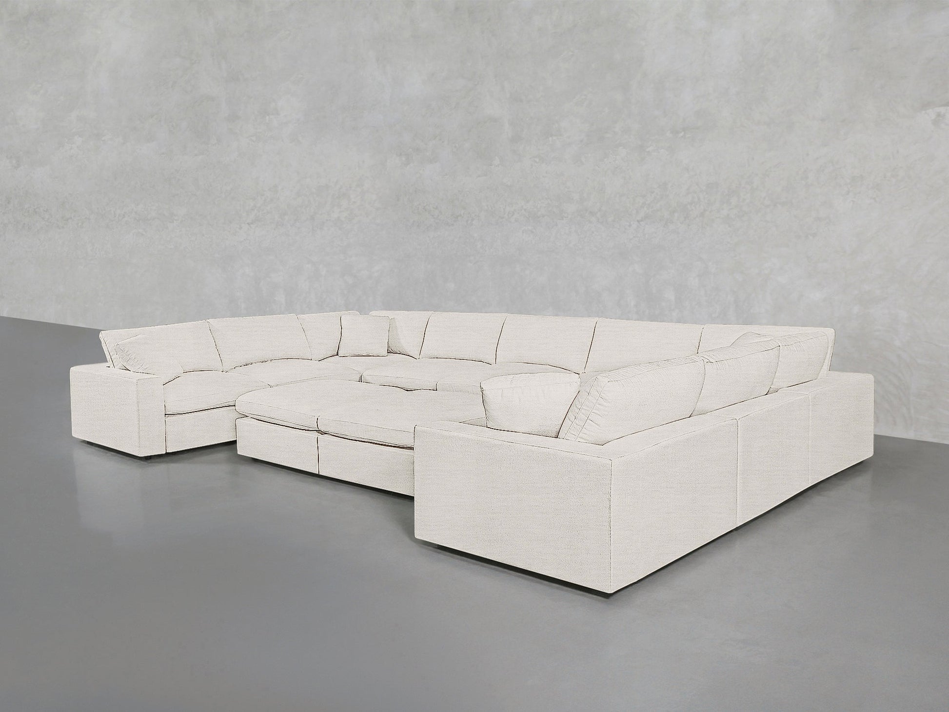 The 9-Seat Modular U-Sectional with Double Ottoman in light beige features wide armrests and plush cushions, arranged on a smooth gray floor against a textured pale gray wall.