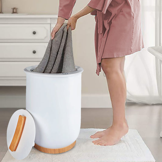 A person in a pink robe places gray towels into the Luxury Bucket Towel Warmer, a large white warmer for bathrooms that fits up to 2 oversized towels, while standing barefoot on a bathroom rug.