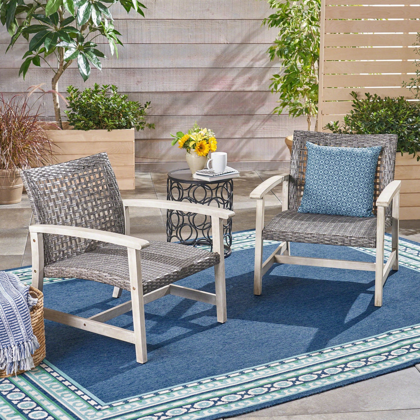 Two gray outdoor club chairs with rattan covers and acacia wood frames sit on a blue patterned rug. A small round table with a mug, flowers, and a book is between them, with plants and a privacy screen behind.