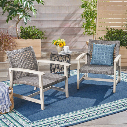 Two gray outdoor club chairs with rattan covers and acacia wood frames sit on a blue patterned rug. A small round table with a mug, flowers, and a book is between them, with plants and a privacy screen behind.