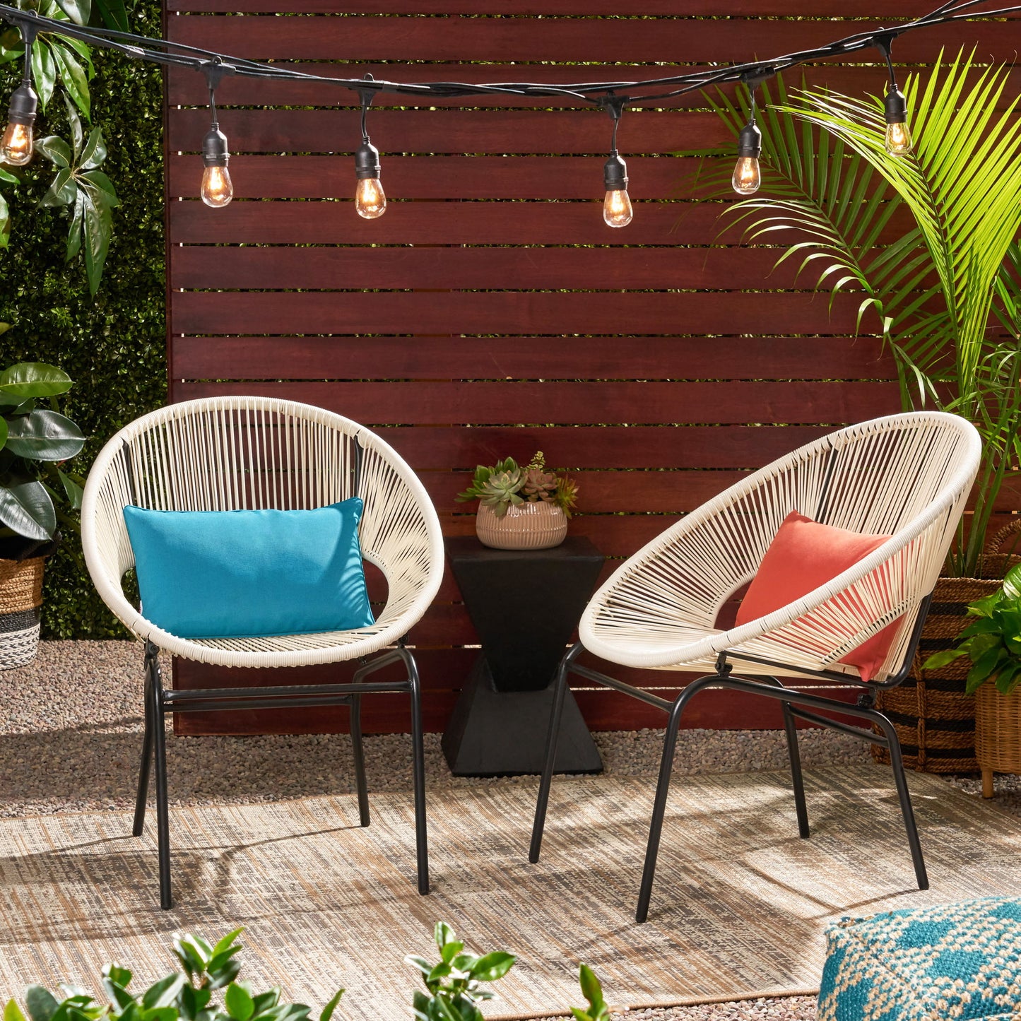 Two Nusa Modern Chair Outdoor Seating pieces with woven seats—one with a blue pillow, one with a coral pillow—sit on an outdoor rug. String lights glow above, surrounded by plants and a wooden privacy fence.