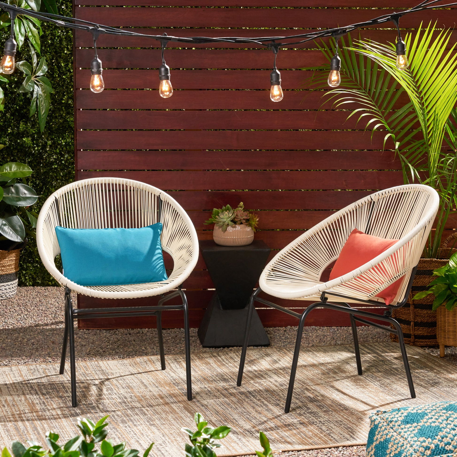 Two Nusa Modern Chair Outdoor Seating pieces with woven seats—one with a blue pillow, one with a coral pillow—sit on an outdoor rug. String lights glow above, surrounded by plants and a wooden privacy fence.