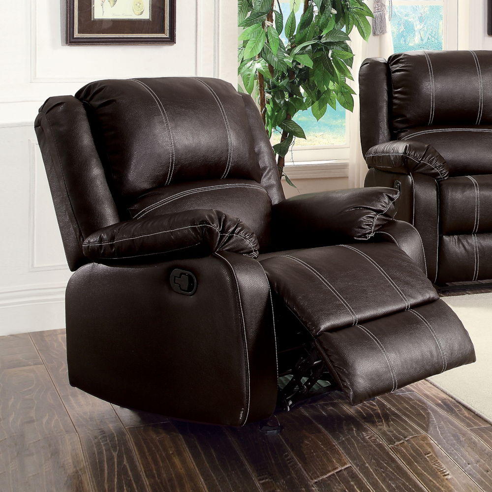 The Zuriel Synthetic Leather Recliner Chair, in dark brown with padded arms and backrest, sits on a wooden floor in a bright living room beside a matching sofa and a green leafy plant.