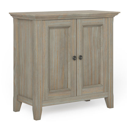 The Amherst - Handcrafted Low Storage Cabinet features a light, weathered wood finish, two paneled doors with round metal knobs, and short, angled legs.