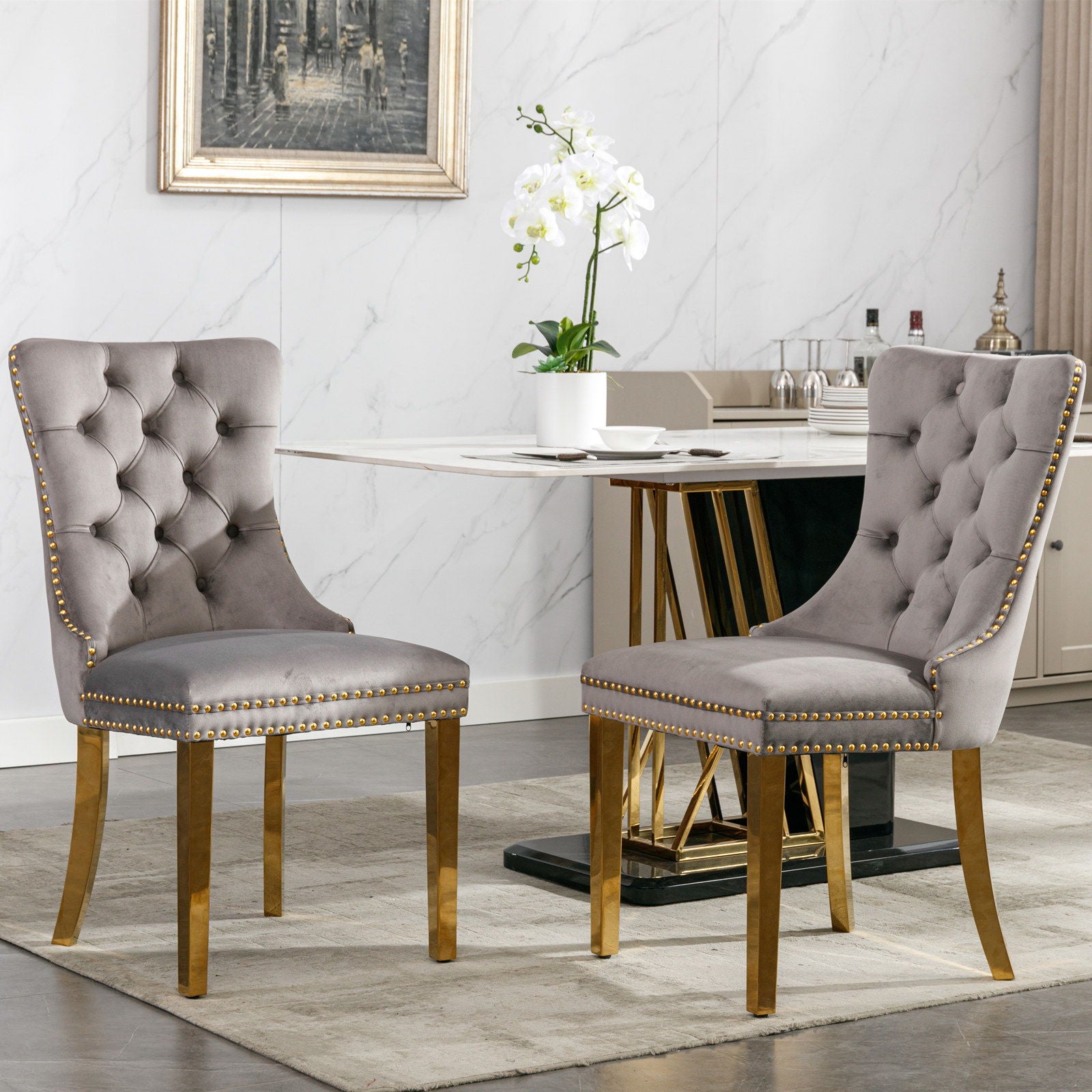 Two Nikki modern tufted velvet dining chairs with golden stainless steel legs and nailhead trim sit by a white marble table, which features a white orchid centerpiece. Framed wall art decorates the background.