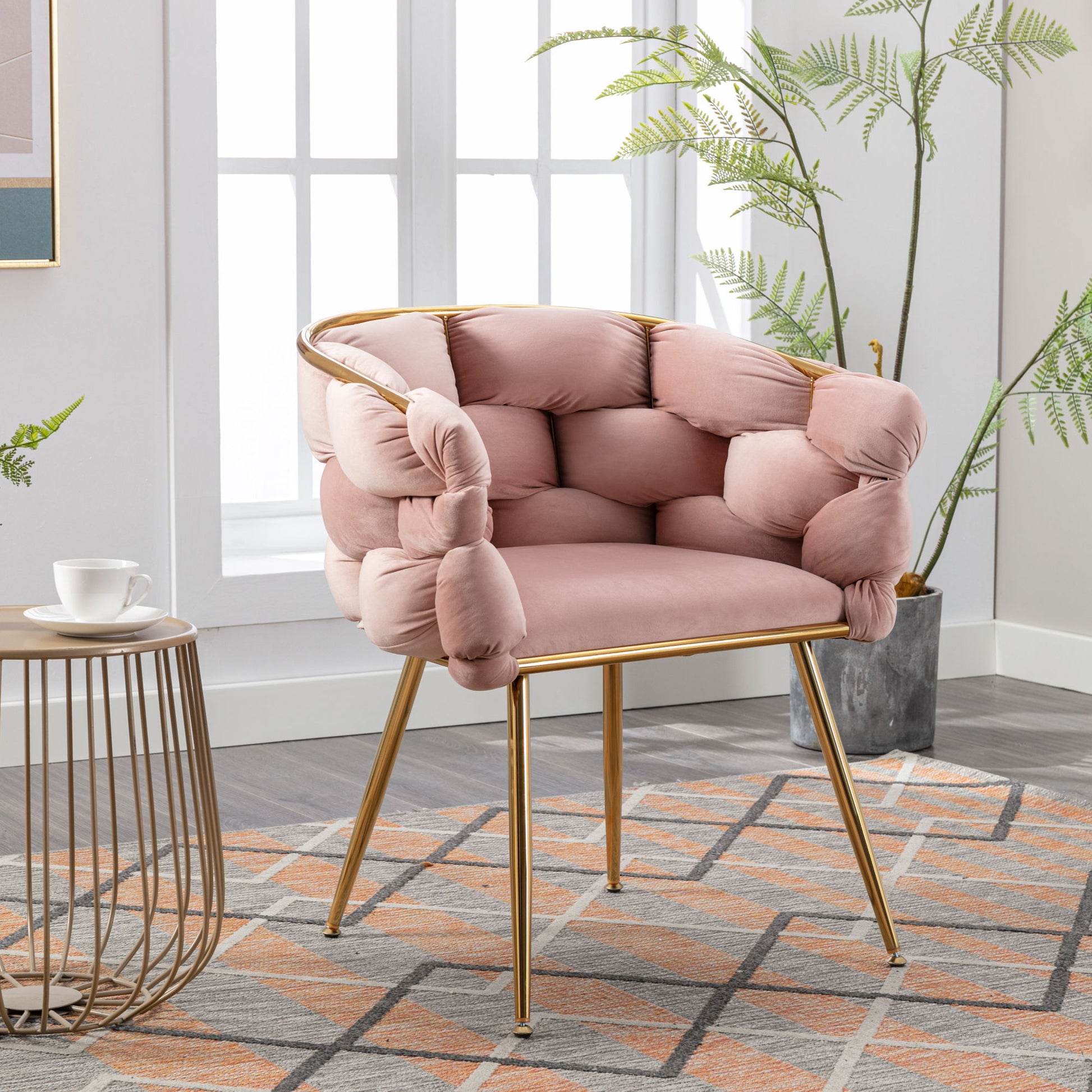 The Luxury Modern Simple Leisure Velvet Single Sofa Chair with tufted pink upholstery and gold legs sits on a geometric rug near a small round table with a teacup in a bright room filled with plants and large windows.