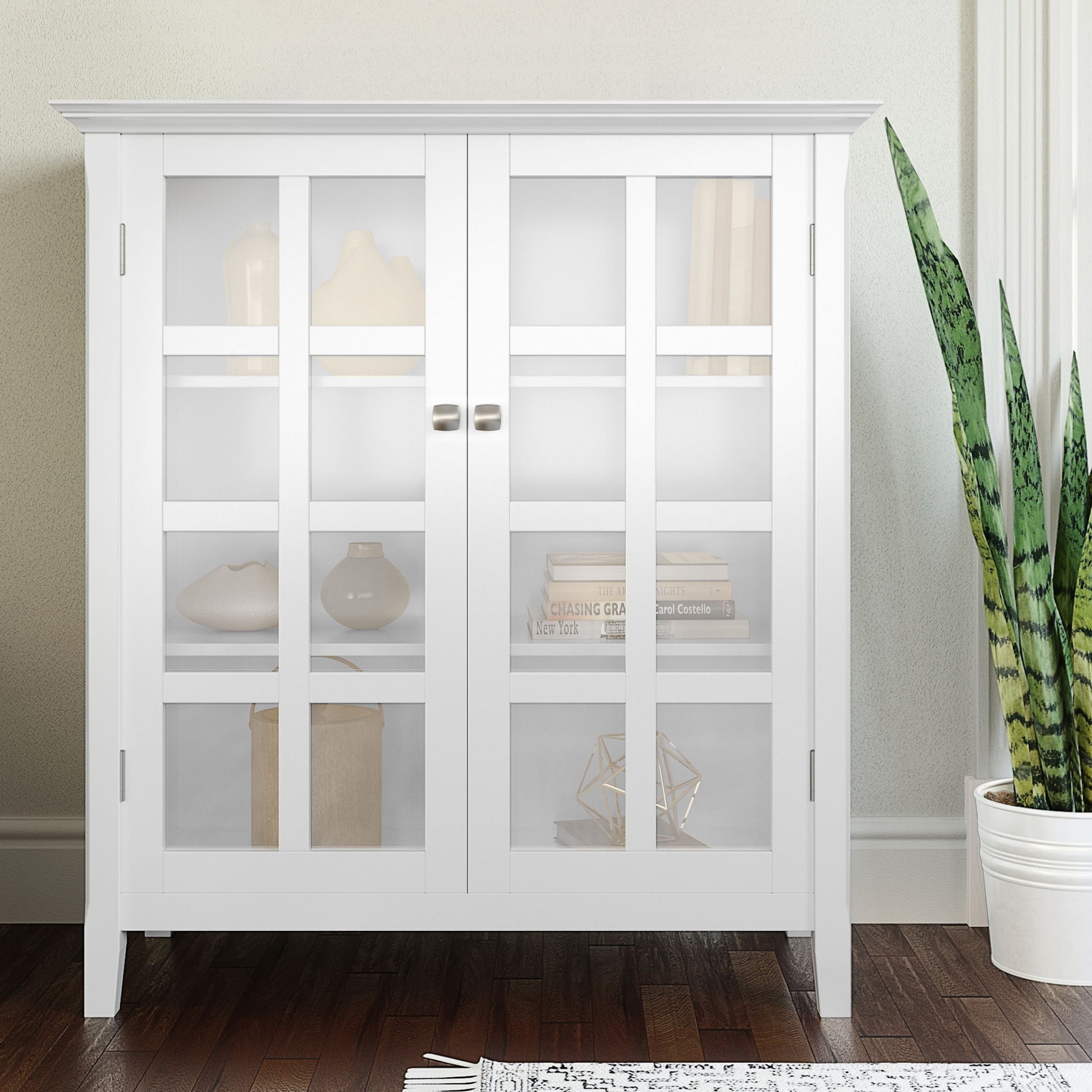 The Acadian Medium Storage Cabinet, handcrafted in white with glass doors, displays neutral vases and books. A green snake plant stands to the right. The cabinet sits on a dark wood floor against a light wall.