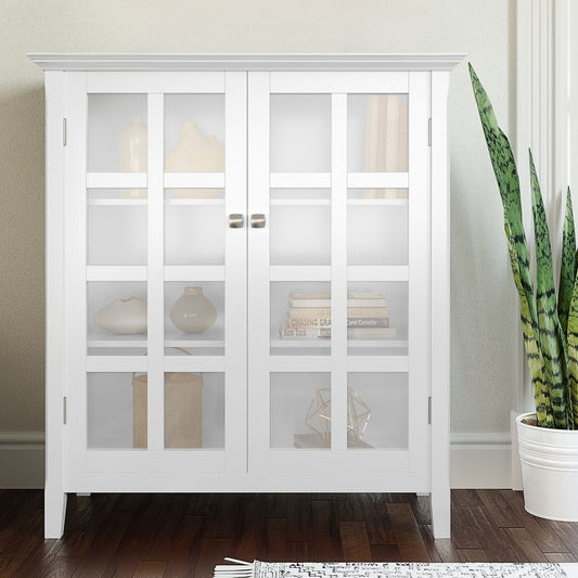 The Acadian Medium Storage Cabinet, handcrafted in white with glass doors, displays neutral vases and books. A green snake plant stands to the right. The cabinet sits on a dark wood floor against a light wall.
