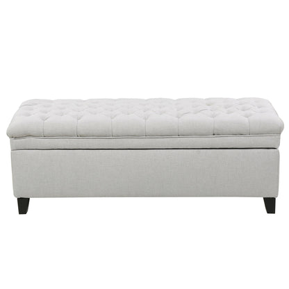 The Juliana Storage Ottoman features a light gray tufted cushioned top and dark birch legs, displayed against a plain white background.