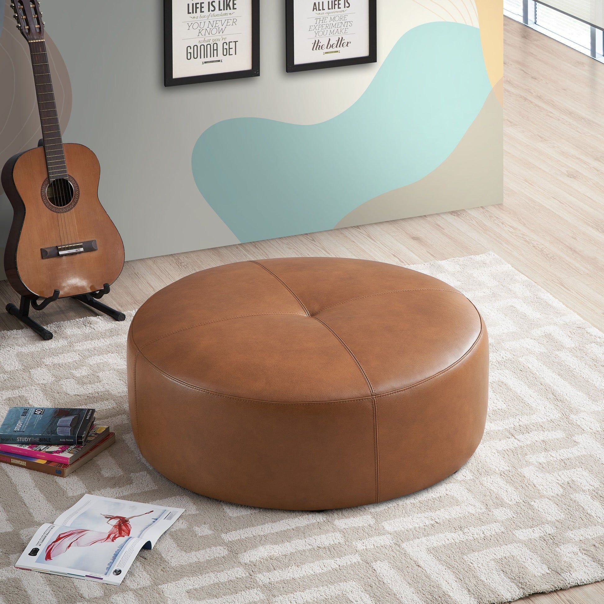 The Rochelin - Ottoman - Tan, large and round, is on a patterned rug beside an acoustic guitar, magazines, and a sketchbook; abstract wall art and light wood flooring complete the scene.