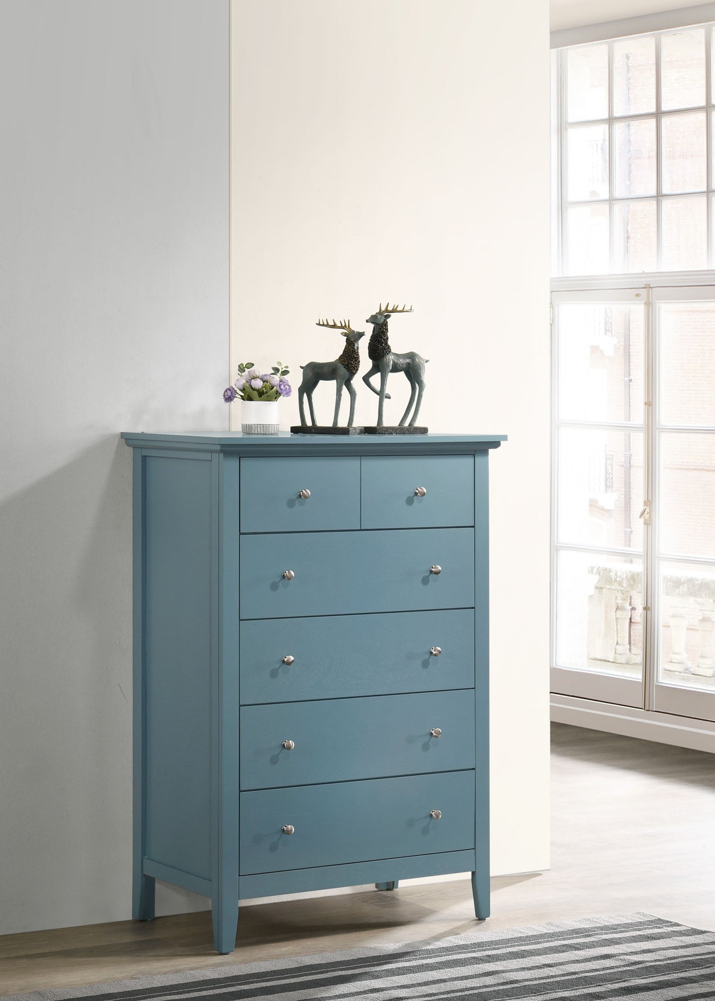 The Hammond Chest, featuring a blue finish and silver knobs, stands by a large window. On its top are two moose figurines and a small potted plant, all illuminated by sunlight in the minimalist room.