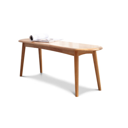 The Dining Bench, Table Bench For Living Room features four angled legs, rounded corners, and a smooth finish. An open magazine sits on top of the bench against a plain white background.