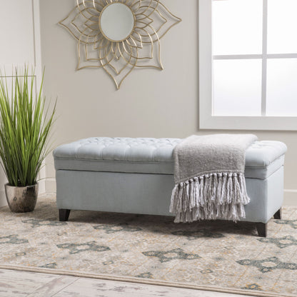 The Juliana Storage Ottoman With Birch Legs in light blue sits on a patterned rug beside a tall potted plant, topped with a gray fringed throw. Above, a round decorative mirror hangs on the wall as sunlight streams through the window.