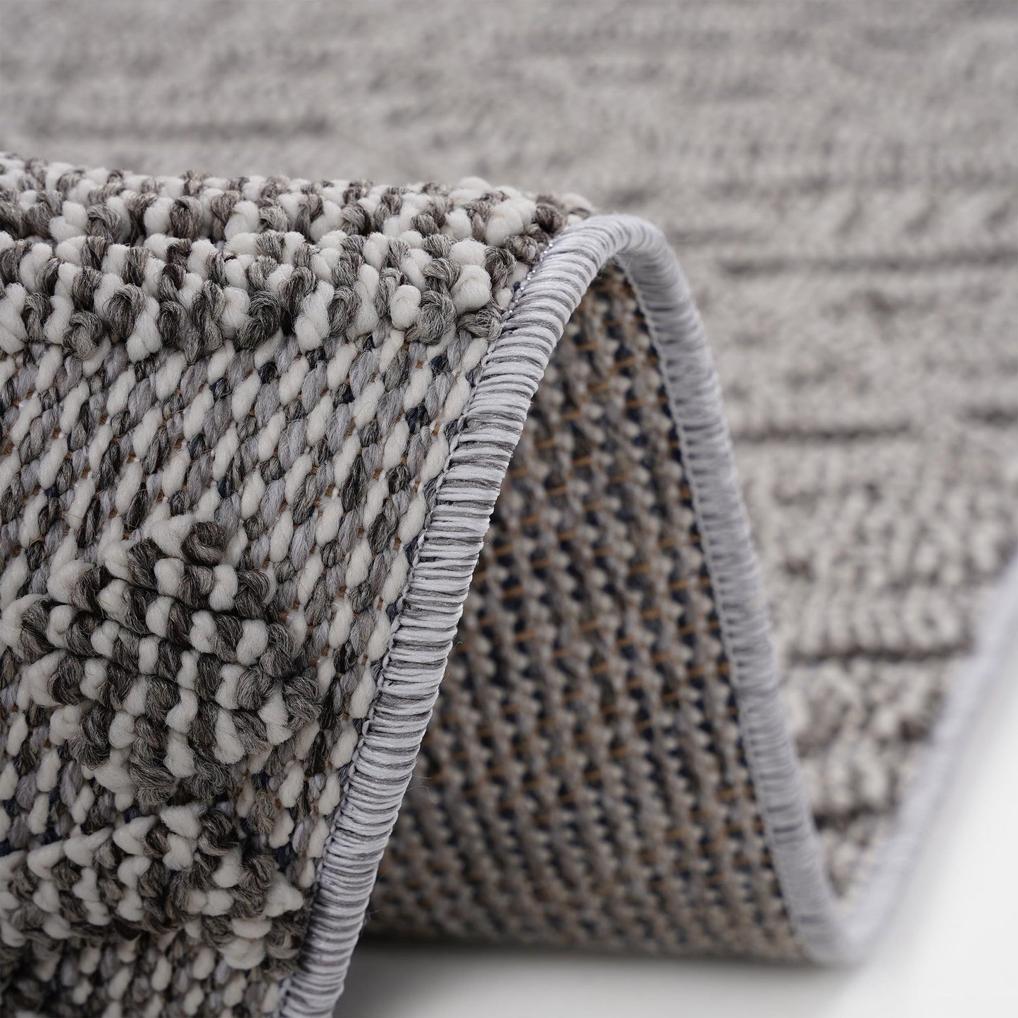 Close-up of the Guros - Traditional Indoor / Outdoor Area Rug in gray and white, highlighting its textured woven pattern with a curled edge that displays both the surface design and underside stitching.