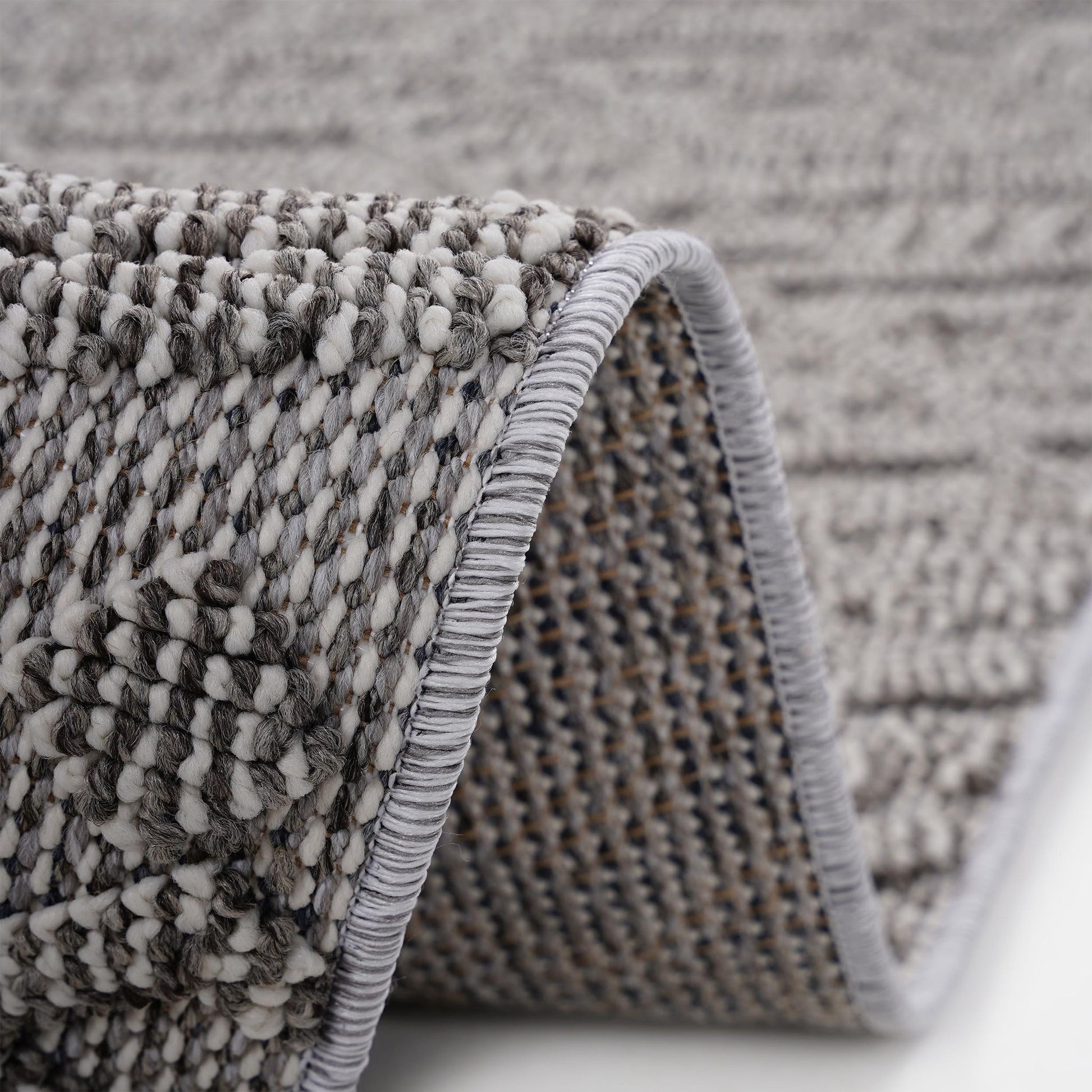 Close-up of the Guros - Traditional Indoor / Outdoor Area Rug in gray and white, highlighting its textured woven pattern with a curled edge that displays both the surface design and underside stitching.