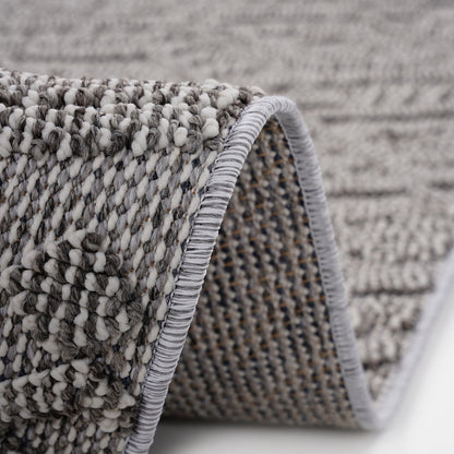 Close-up of the Guros - Traditional Indoor / Outdoor Area Rug in gray and white, highlighting its textured woven pattern with a curled edge that displays both the surface design and underside stitching.