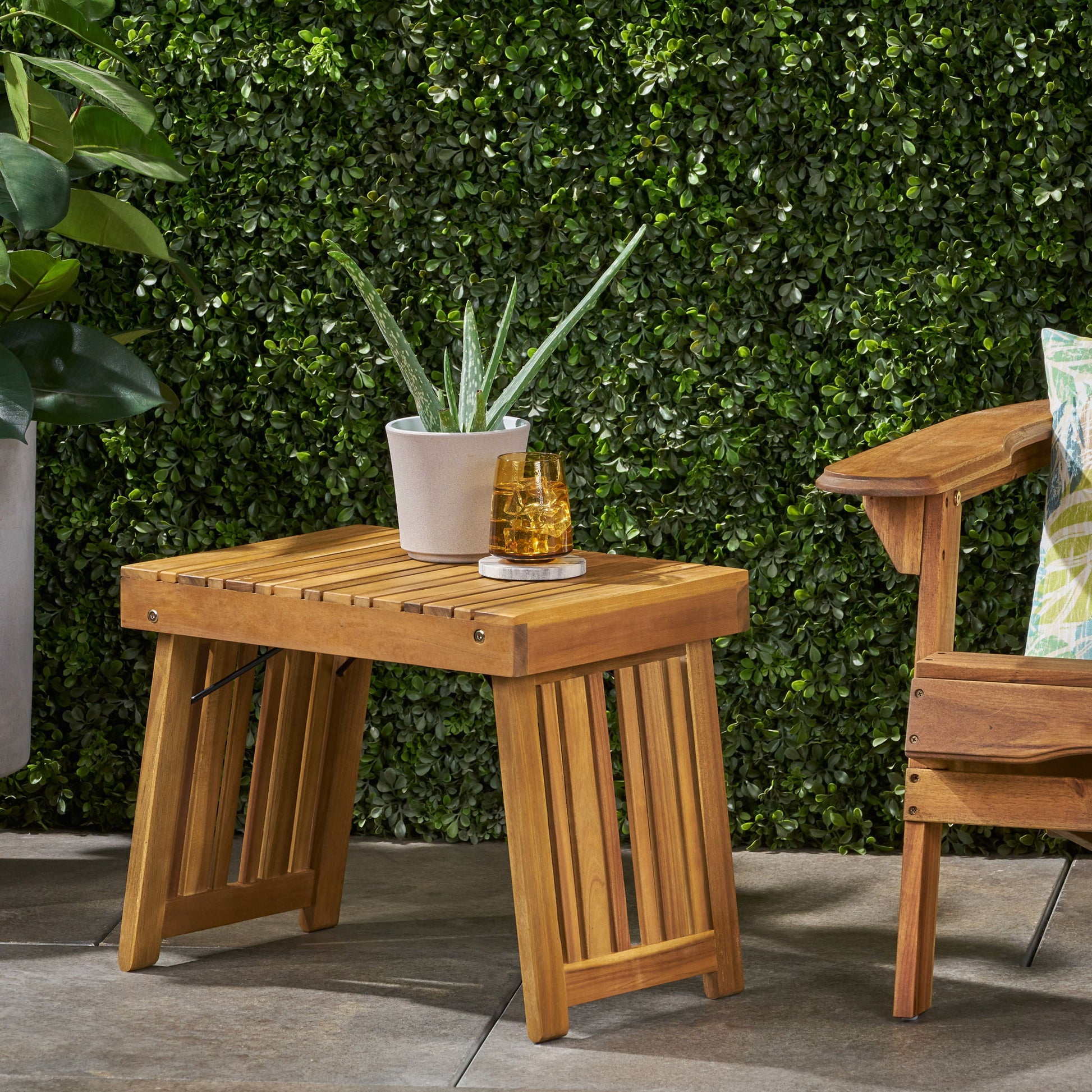The Temptation Outdoor Foldable Side Table with slat design sits on a patio, holding a potted aloe and two amber glasses, beside a wooden chair with a green patterned cushion and lush foliage in the background.