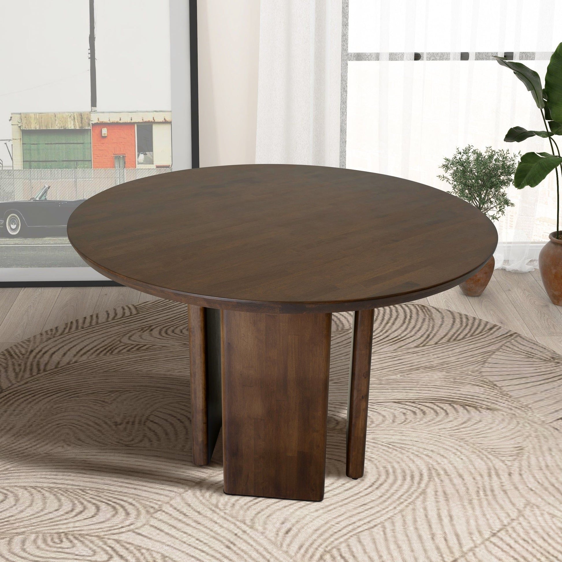 The Alden Round Dining Table in walnut features a unique intersecting base and sits on a beige patterned rug in a modern room with a framed photo, potted plants, and sheer curtains in the background.