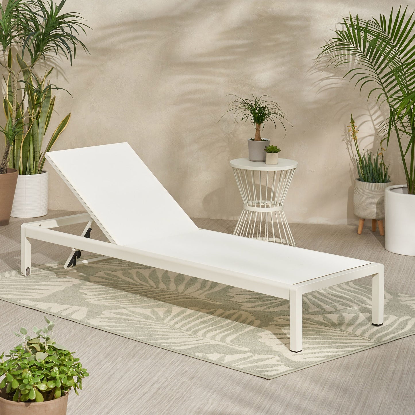 The Outdoor Patio Chaise Lounge With 5 Reclining Positions, finished in modern white, rests on a patterned rug among potted green plants beside a small round side table with a plant, all set against a light-colored wall.