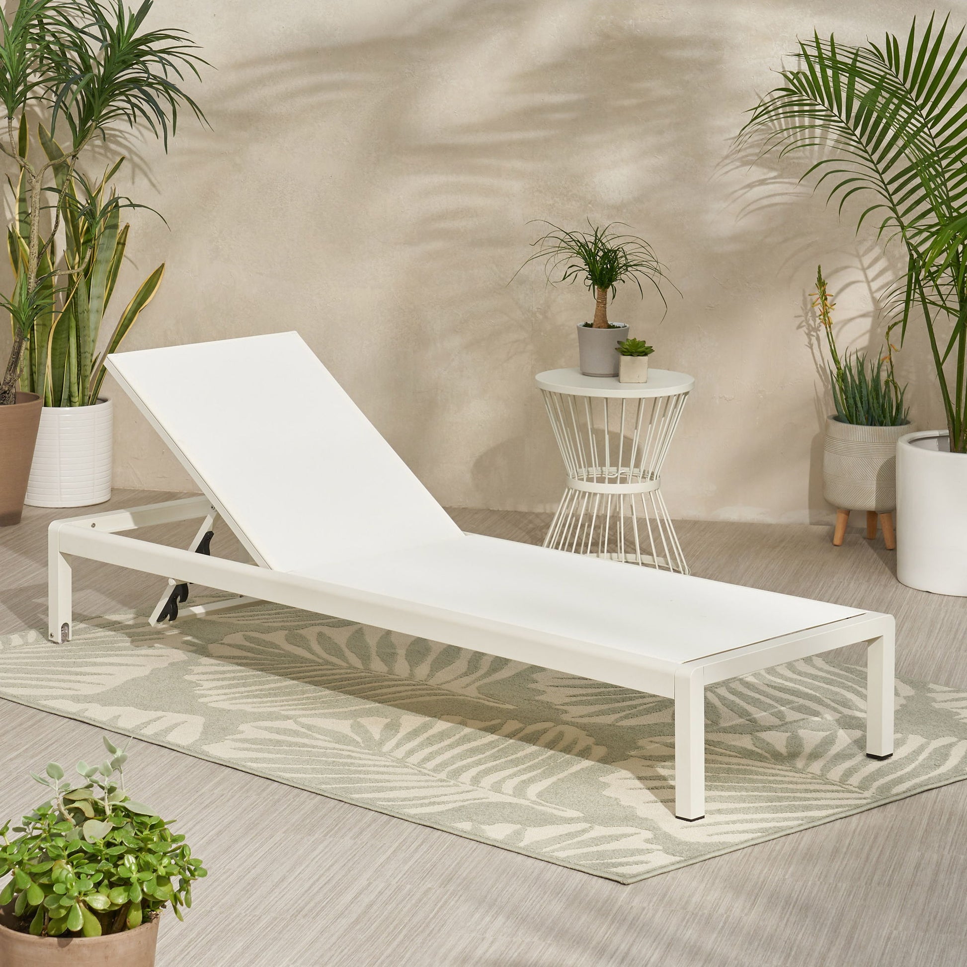 The Outdoor Patio Chaise Lounge With 5 Reclining Positions, finished in modern white, rests on a patterned rug among potted green plants beside a small round side table with a plant, all set against a light-colored wall.