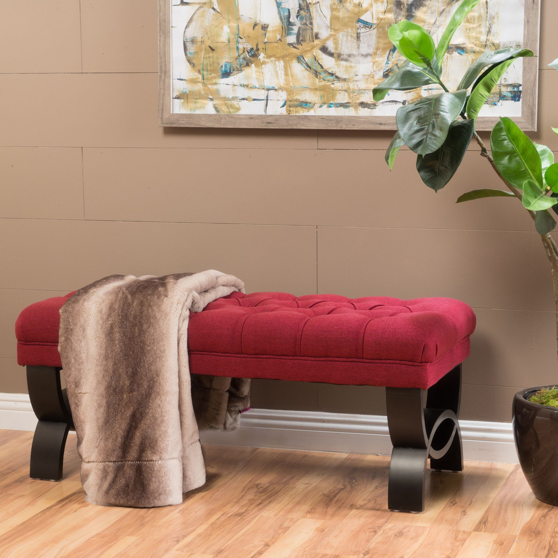 The Hilton Ottoman Bench Classic Style with red tufted upholstery and dark curved legs sits on a wooden floor, draped with a brown faux fur blanket. A green potted plant is nearby, and an abstract painting hangs on the tan wall.