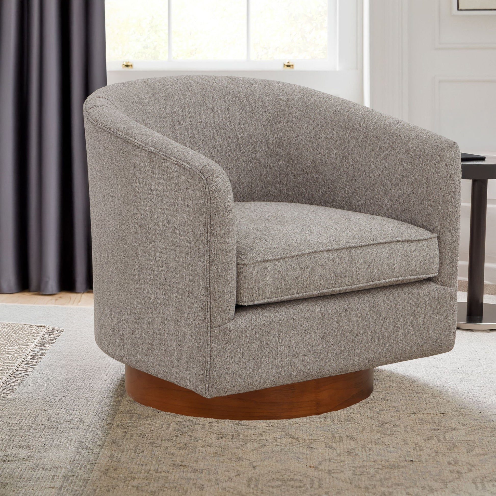 The Miramar Swivel Barrel Chair with a wooden base and modern gray upholstery sits on a light patterned rug in a bright, stylish living room.