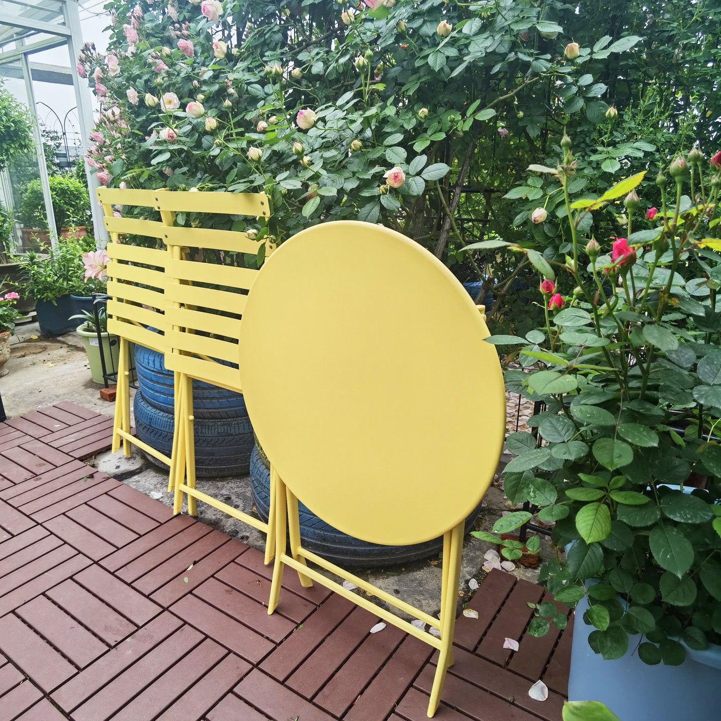 The 3 Piece Patio Bistro Set of a foldable round table and two chairs is arranged on a red-tiled patio, surrounded by lush green rose bushes blooming with pink flowers.
