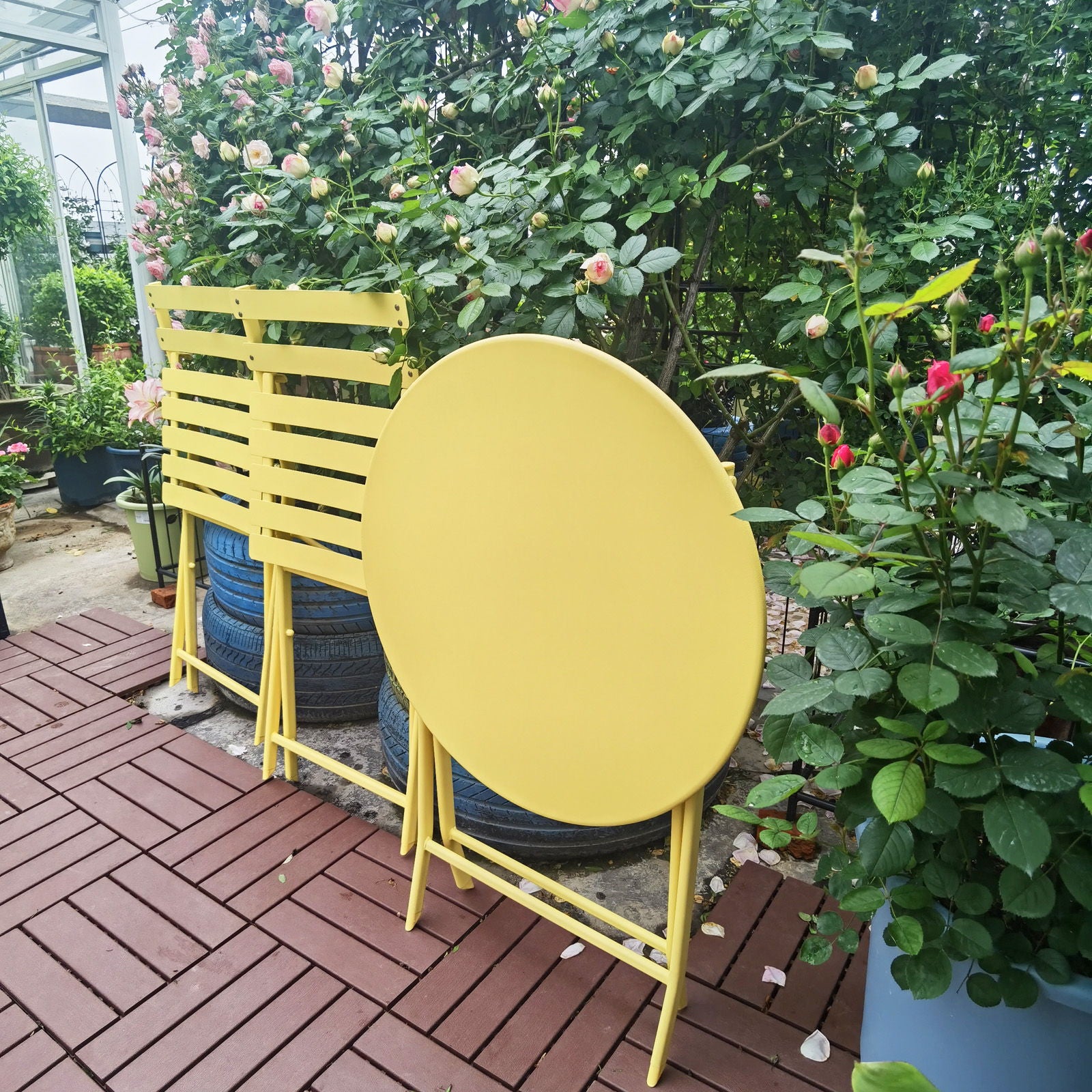 The 3 Piece Patio Bistro Set of a foldable round table and two chairs is arranged on a red-tiled patio, surrounded by lush green rose bushes blooming with pink flowers.