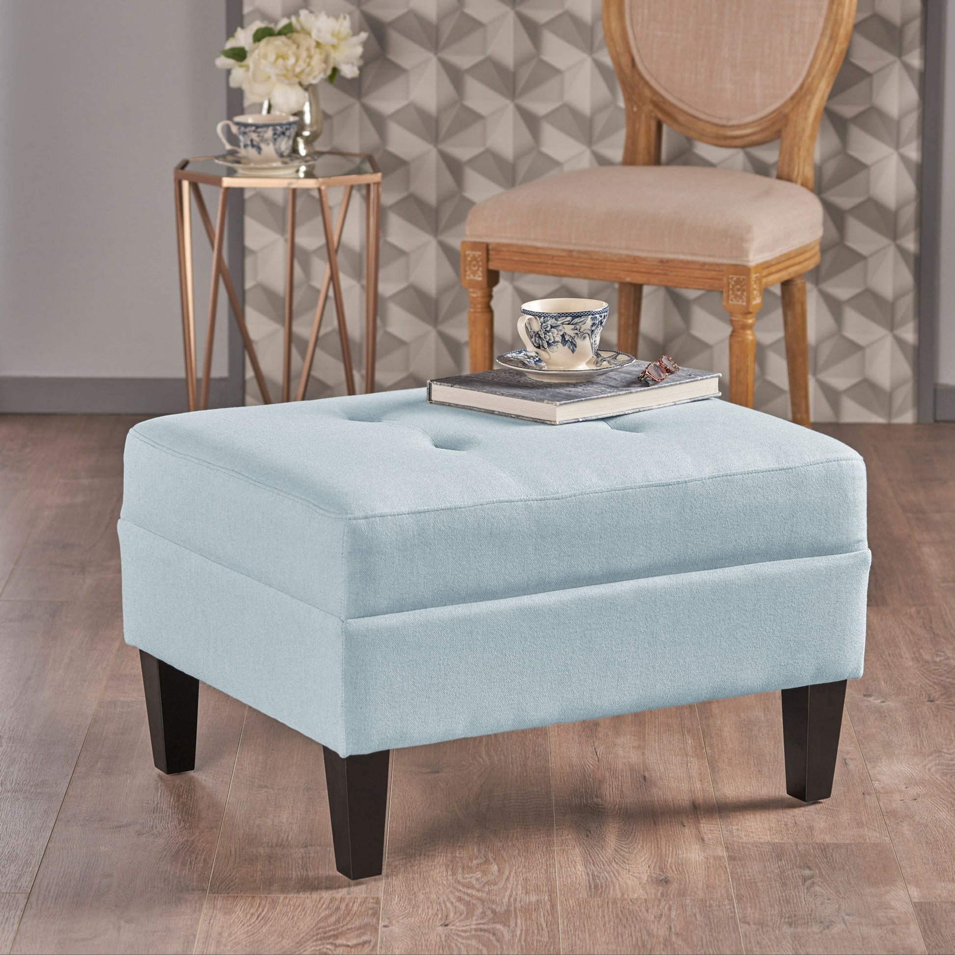 A Classic Ottoman With Birch Wood Legs And Fabric Upholstery in light blue sits on a wooden floor with a book and teacup on top. A beige chair, flower-topped table, and patterned wallpaper are in the background.