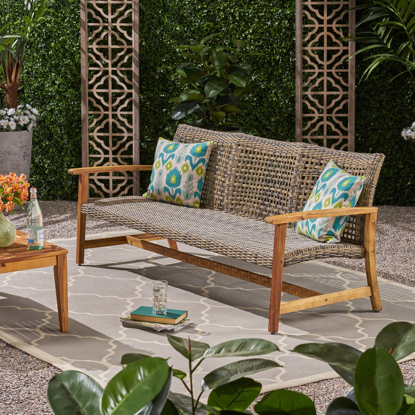 A gray Outdoor 3 Seater Sofa With Rattan Cover featuring two patterned pillows is placed on a rug in a garden, surrounded by lush greenery. A wooden table holding flowers, books, and drinks stands nearby.