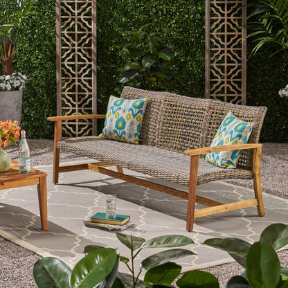 A gray Outdoor 3 Seater Sofa With Rattan Cover featuring two patterned pillows is placed on a rug in a garden, surrounded by lush greenery. A wooden table holding flowers, books, and drinks stands nearby.