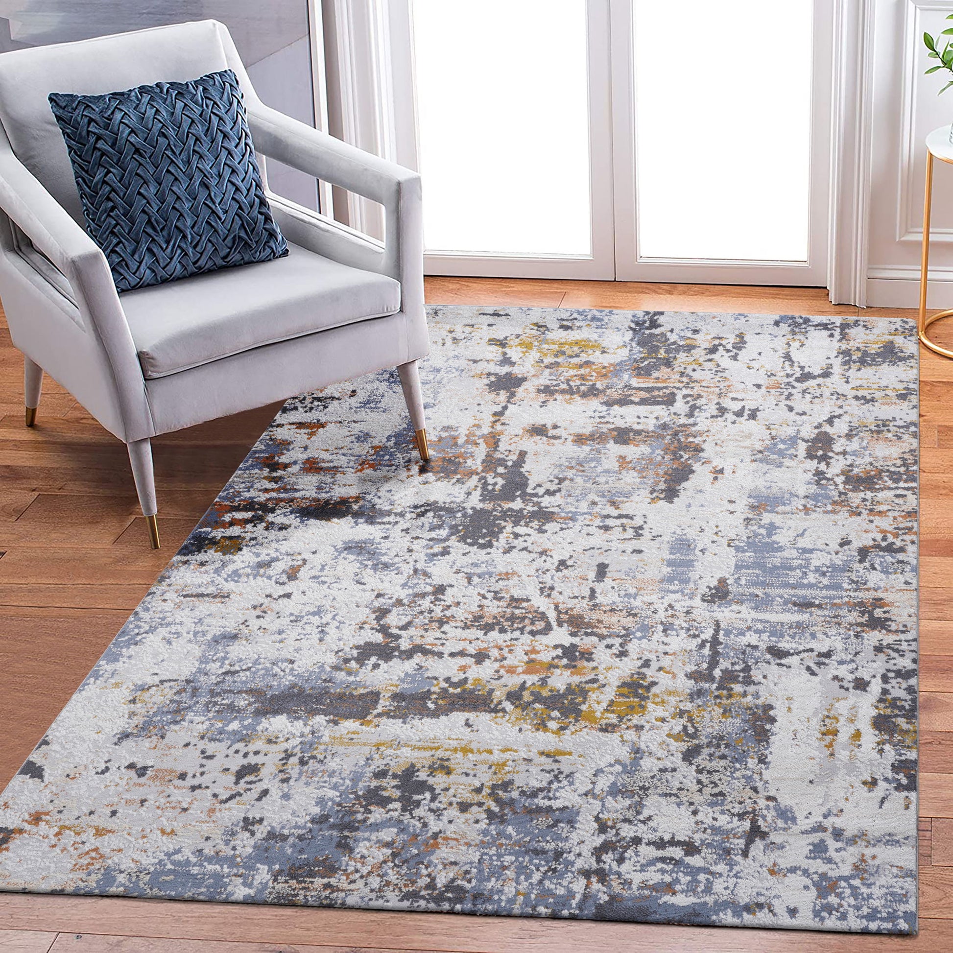 A modern living room features a light gray armchair with a blue patterned pillow on the Payas - Abstract Area Rug, showcasing shades of gray, blue, yellow, and white atop a hardwood floor near glass doors.