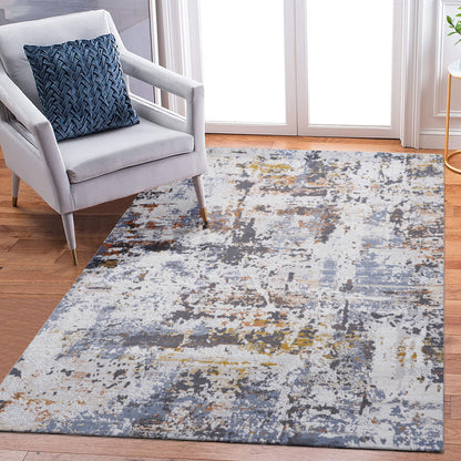 A modern living room features a light gray armchair with a blue patterned pillow on the Payas - Abstract Area Rug, showcasing shades of gray, blue, yellow, and white atop a hardwood floor near glass doors.