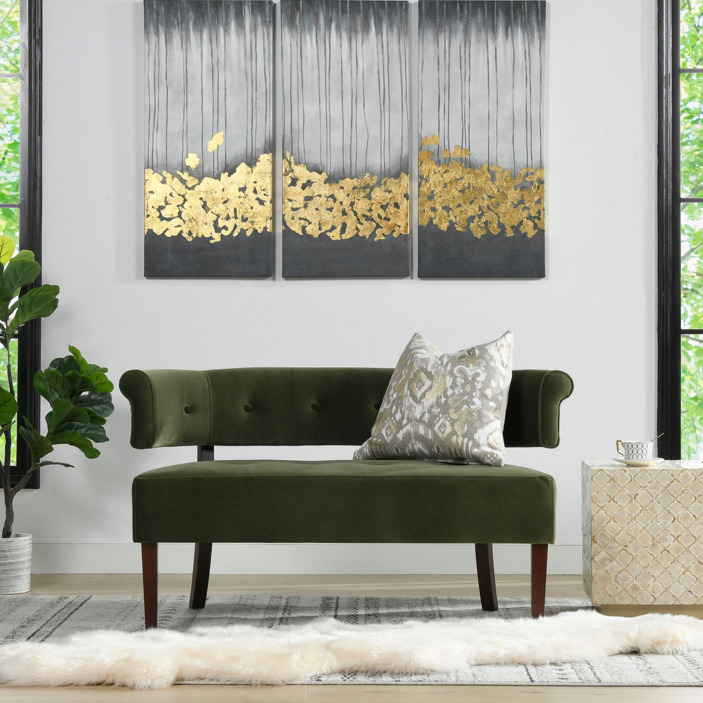 A modern living room with the Jared - Versatile Roll Arm Tufted Bench Settee in green velvet, a patterned throw pillow, abstract gold and gray triptych wall art, large windows, a potted plant, and a light rug topped with a fur accent.