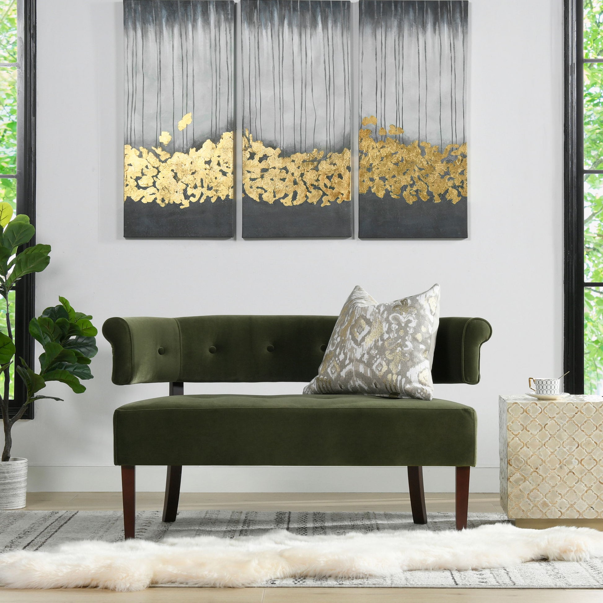 A modern living room with the Jared - Versatile Roll Arm Tufted Bench Settee in green velvet, a patterned throw pillow, abstract gold and gray triptych wall art, large windows, a potted plant, and a light rug topped with a fur accent.