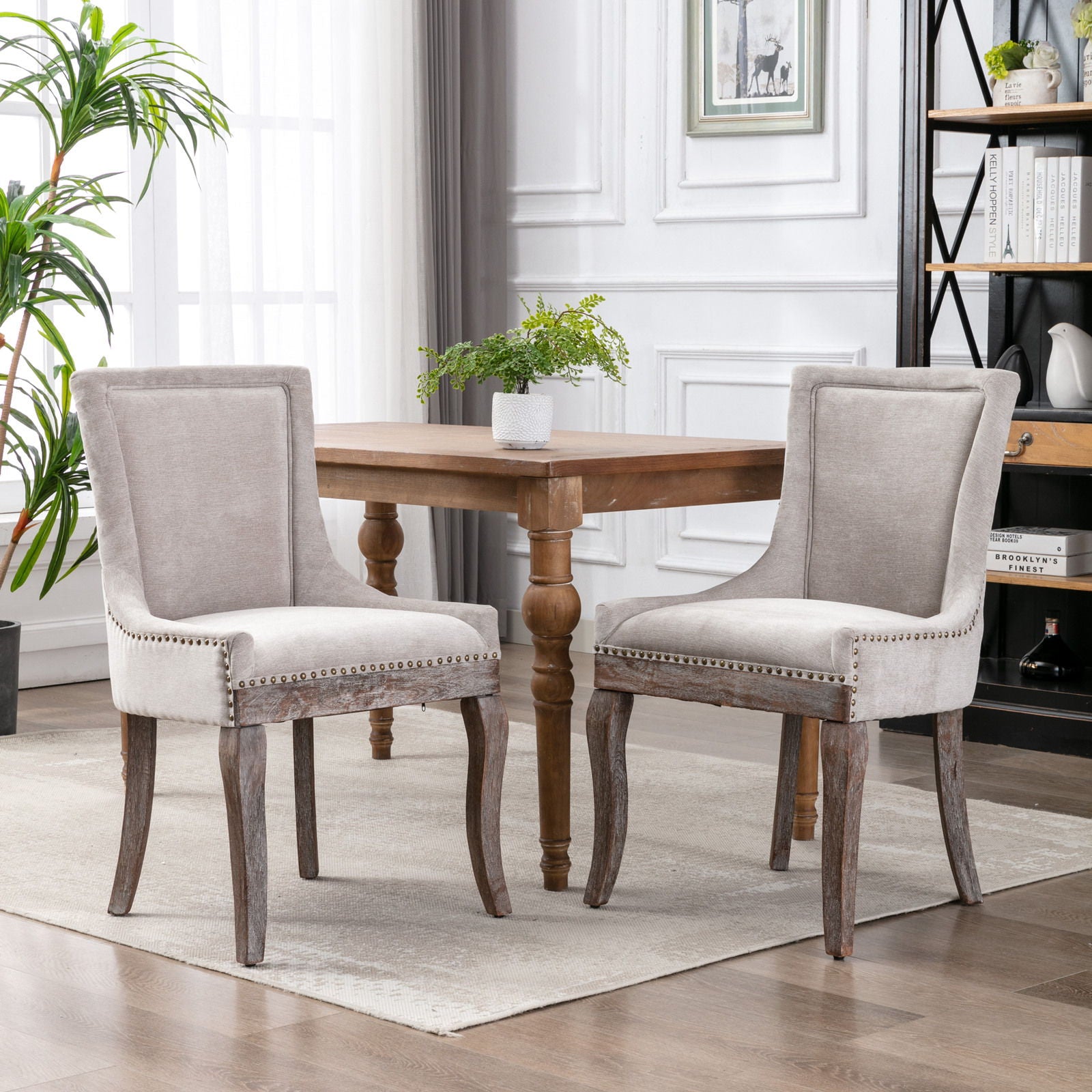 Two Ultra Side Dining Chairs with thickened gray fabric, neutrally toned wood legs, and bronze nail head trim are arranged around a wooden dining table in a bright, modern room with white walls and large windows.