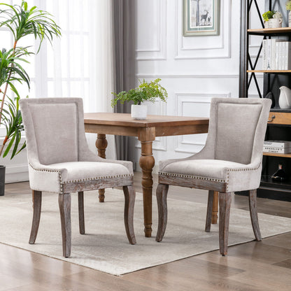 Two Ultra Side Dining Chairs with thickened gray fabric, neutrally toned wood legs, and bronze nail head trim are arranged around a wooden dining table in a bright, modern room with white walls and large windows.