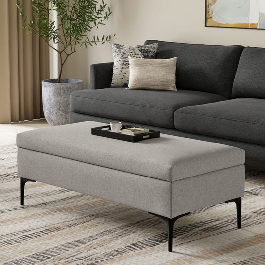 A modern living room features the Rebecca Multifunctional Coffee Table Storage Ottoman with a gray upholstered lift-up lid, black metal legs, and a black tray with a cup and saucer, in front of a dark gray sofa, potted plant, and patterned rug.