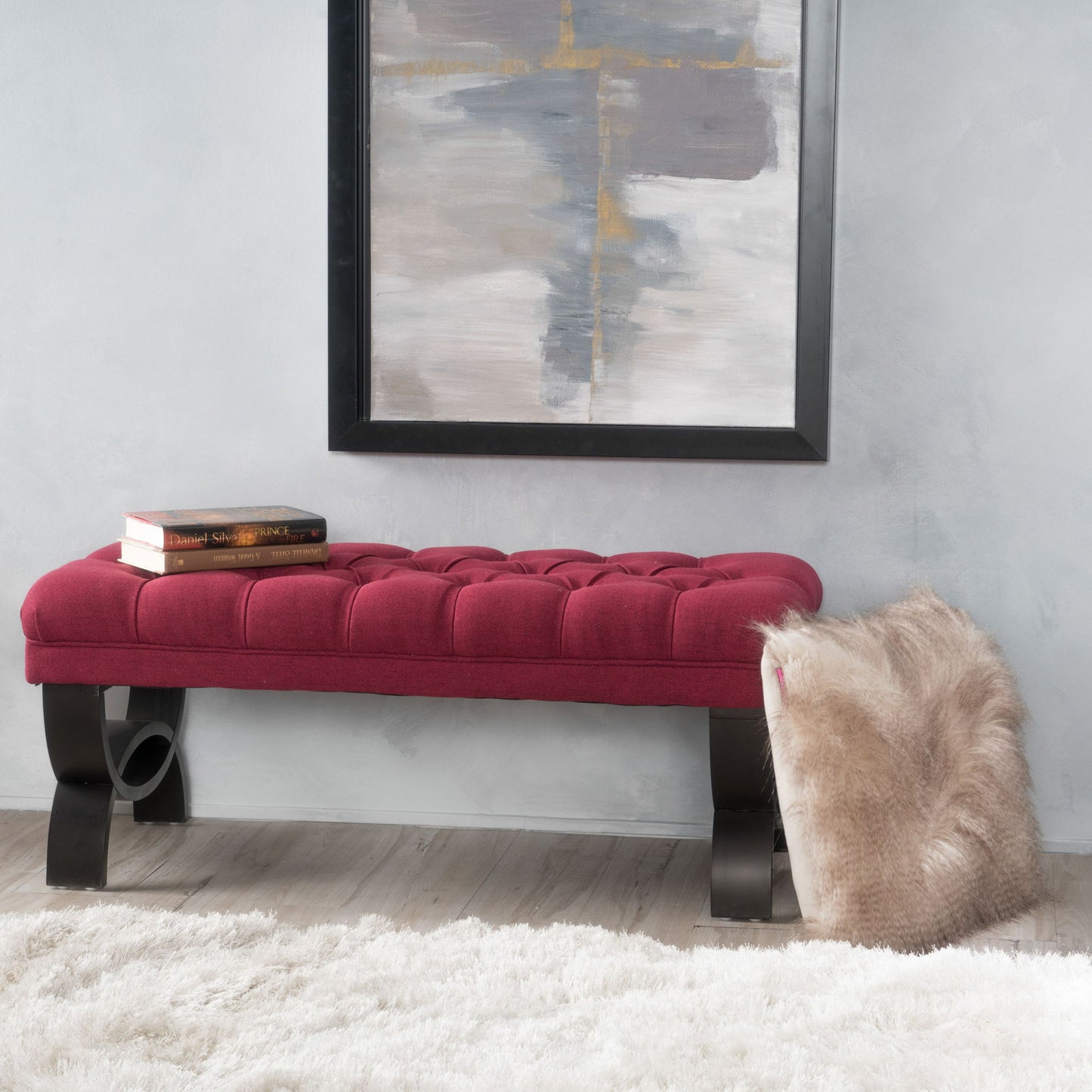The Hilton - Ottoman Bench Classic Style, featuring a red tufted seat and black curved legs, sits in front of a gray wall under an abstract painting. A fur throw is draped over one end, two books rest on the other, and a white rug covers the floor.