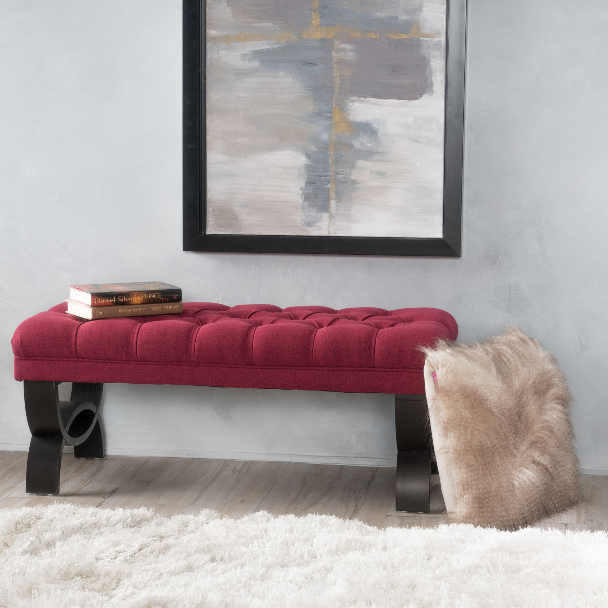 The Hilton - Ottoman Bench Classic Style, featuring a red tufted seat and black curved legs, sits in front of a gray wall under an abstract painting. A fur throw is draped over one end, two books rest on the other, and a white rug covers the floor.