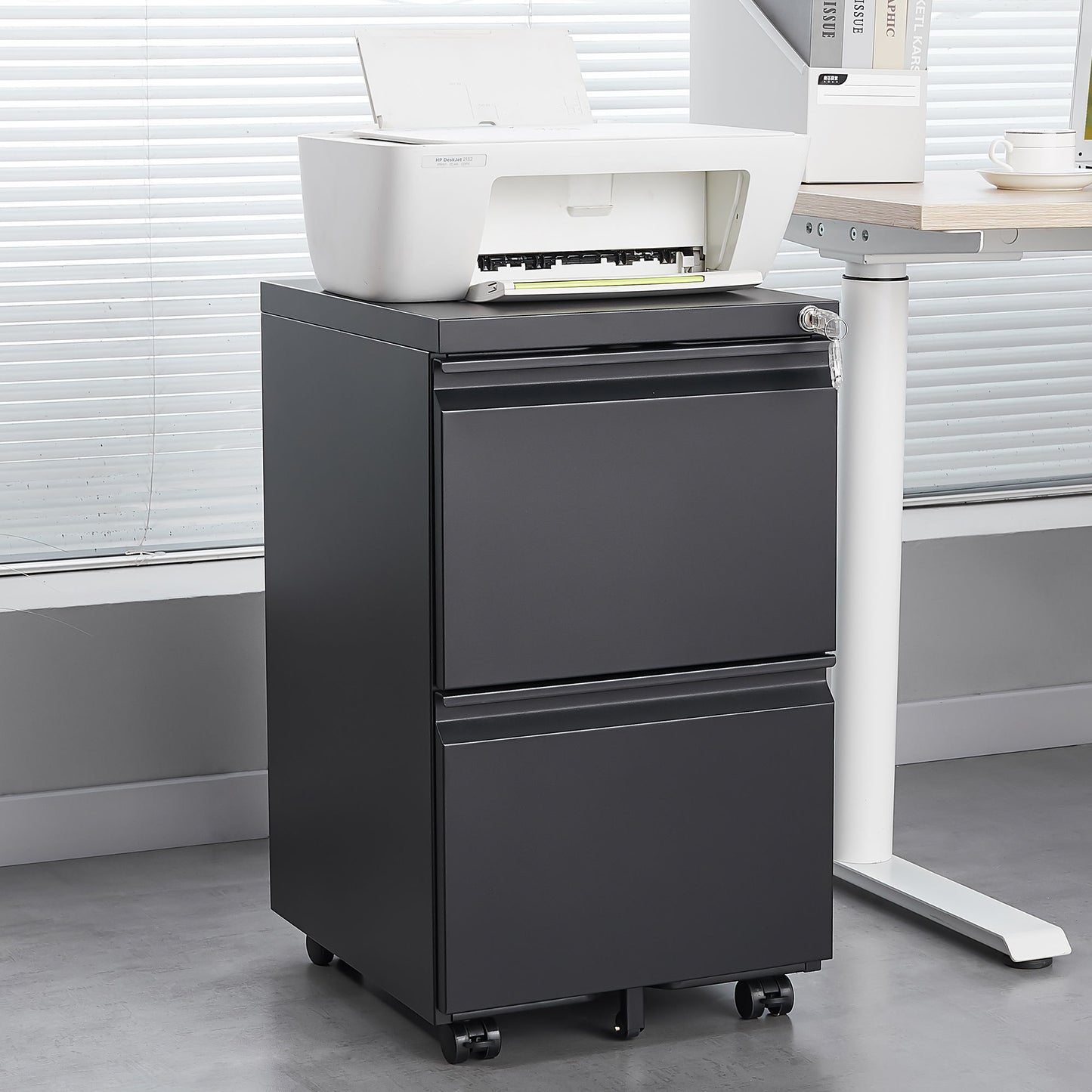 A white printer rests on a 2 Drawer File Cabinet With Lock, a metal under desk rolling filing cabinet for letter/legal/A4 size, next to a desk in a modern office with large window blinds.