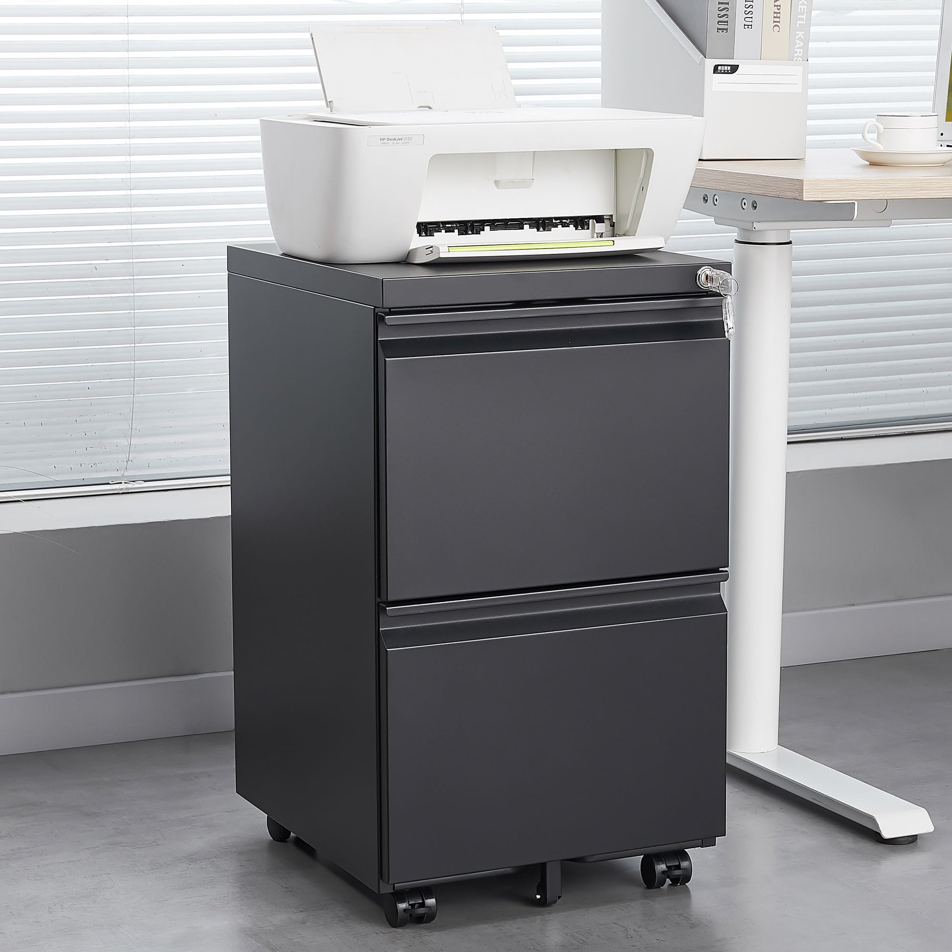 A white printer rests on a 2 Drawer File Cabinet With Lock, a metal under desk rolling filing cabinet for letter/legal/A4 size, next to a desk in a modern office with large window blinds.