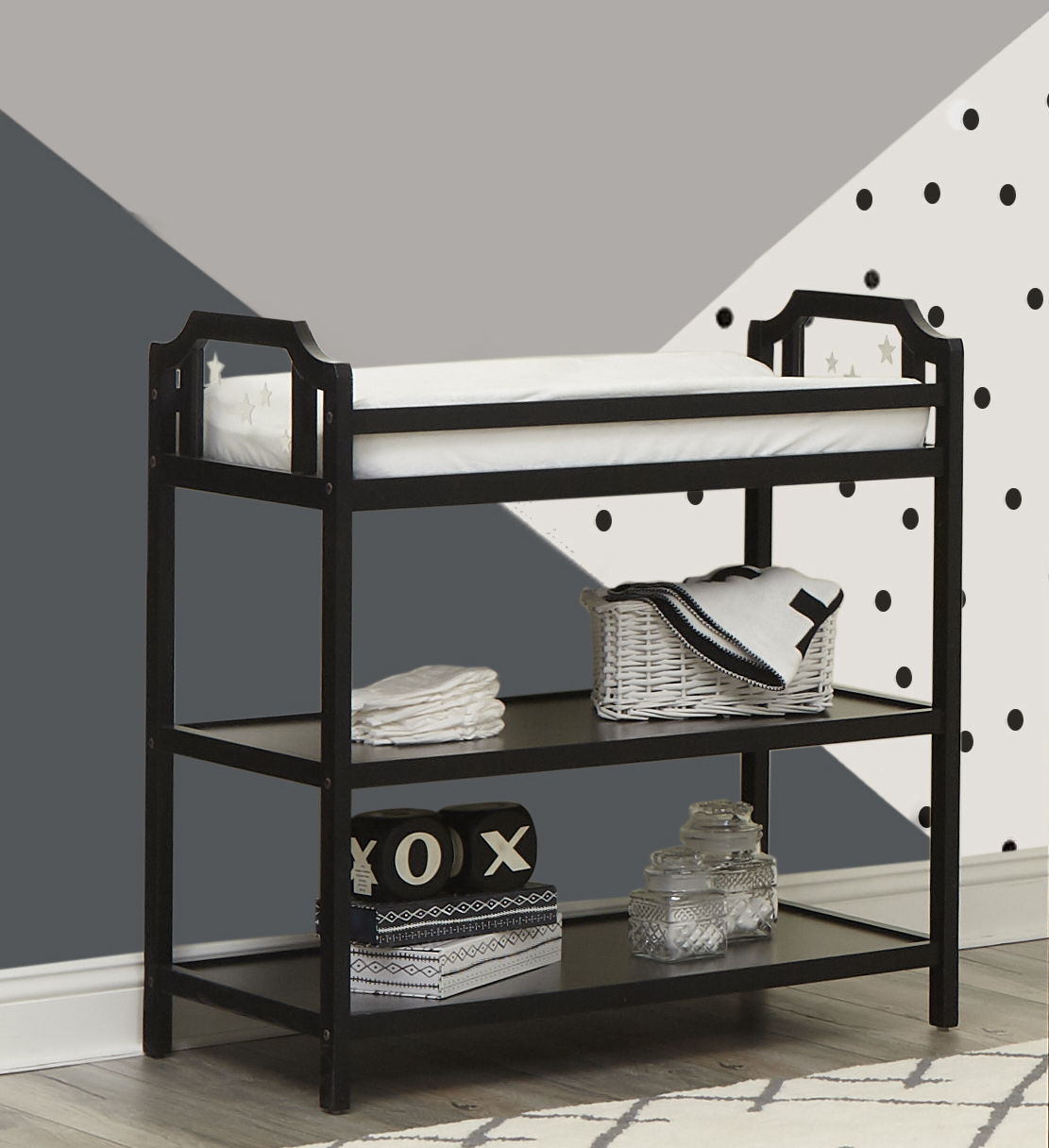 The Celeste Changing Table features a black frame, white cushioned pad, stacked towels, baskets, and glass jars on two shelves, set against a geometric wall with gray, white, and polka dot patterns.