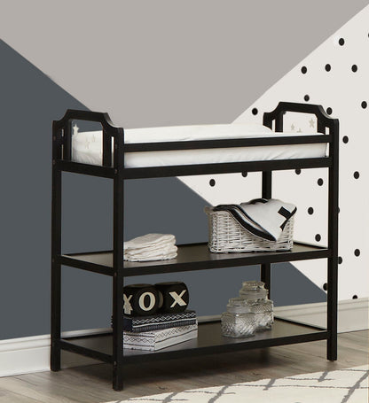 The Celeste Changing Table features a black frame, white cushioned pad, stacked towels, baskets, and glass jars on two shelves, set against a geometric wall with gray, white, and polka dot patterns.